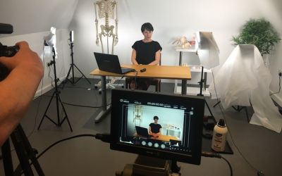 Someone being filmed while sitting at a table, there is a skeleton behind her and cloths covering lights