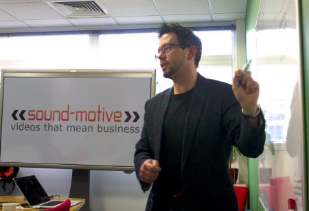 A man presenting, to his left is a screen with "Sound-Motive" "videos that mean business" on it